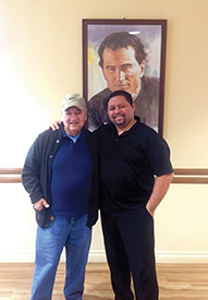 Lucero and Brother Charles Thenier, S.D.B. stand before a portrait of the founder of the Salesians, Saint John Bosco.