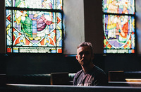 young man sitting in a church pew with stained-glass windows behind